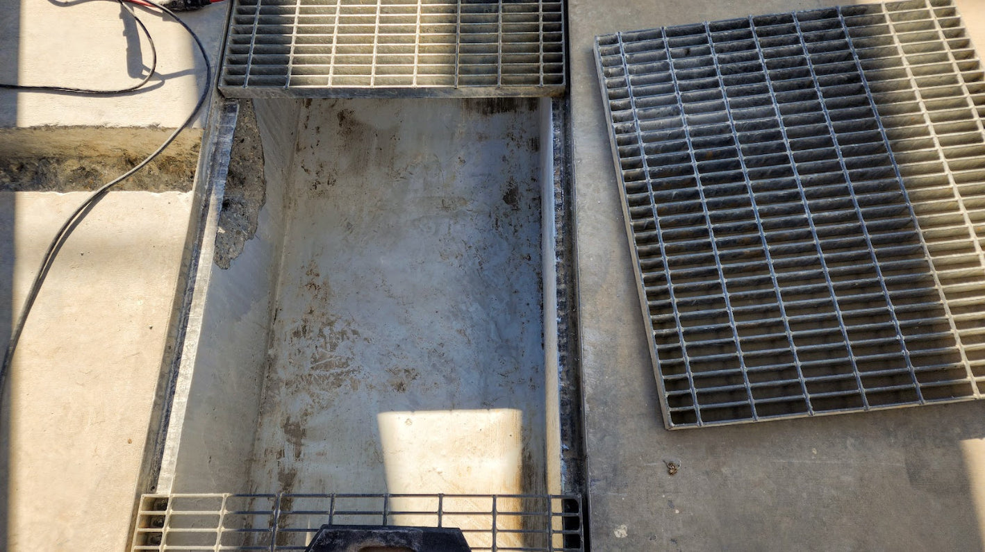 Catch basin with metal grating cover removed, exposing the concrete chamber and drain opening beneath.