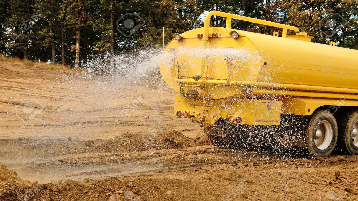 Yellow tanker truck spraying water on a dirt road with trees in the background